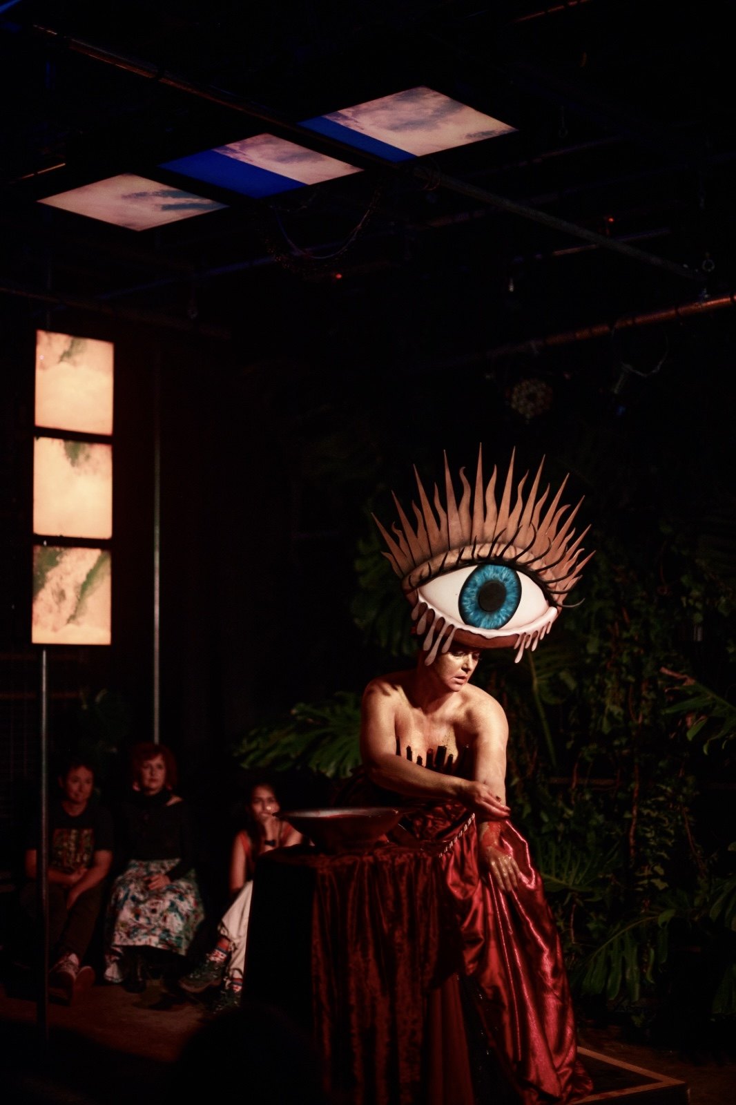 Laura King leaning over a bowl on a red-draped table, wearing the sculpted eye headdress.